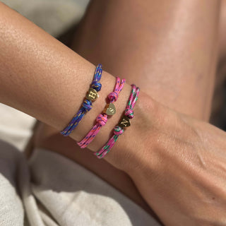 Zwei handgeflochtene Surferarmbänder aus buntem Segeltau mit goldenem Herzanhänger und Initiale, getragen am Handgelenk.