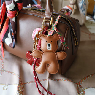 Braune Handtasche mit buntem Skinny-Schal am Griff und einem braunen Teddy-Schlüsselanhänger aus Leder, dekoriert mit farbigen Bändern und Charms.“