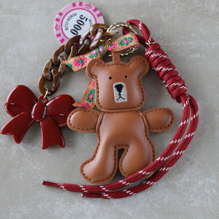 Braune Handtasche mit buntem Skinny-Schal am Griff und einem braunen Teddy-Schlüsselanhänger aus Leder, dekoriert mit farbigen Bändern und Charms.“